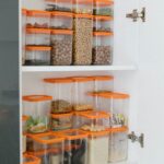 About Neatly organized pantry shelves featuring diverse grains and seeds in transparent containers with orange lids.