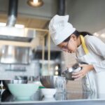 About A young girl in a chef's hat pours ingredients into a mixing bowl in a kitchen.